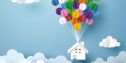 Papieren wit huis hangt aan kleurrijke ballonen met op de achtergrond een blauwe lucht en wolken